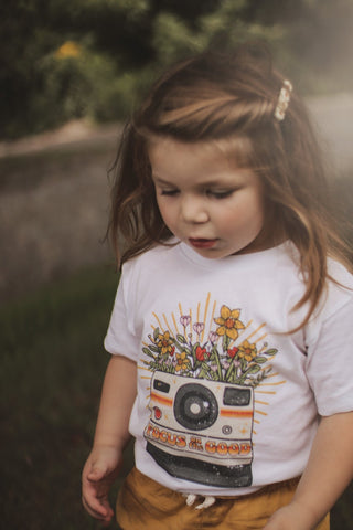 white t-shirt with camera and flowers, reads: focus on the good