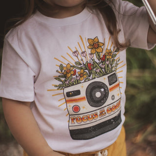 white t-shirt with camera and flowers, reads: focus on the good